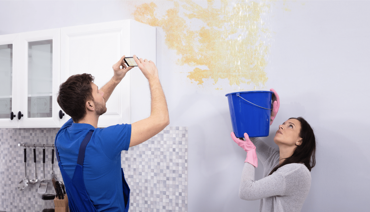 A man photographing a water-stained wall while a woman holds a bucket underneath to catch dripping water.